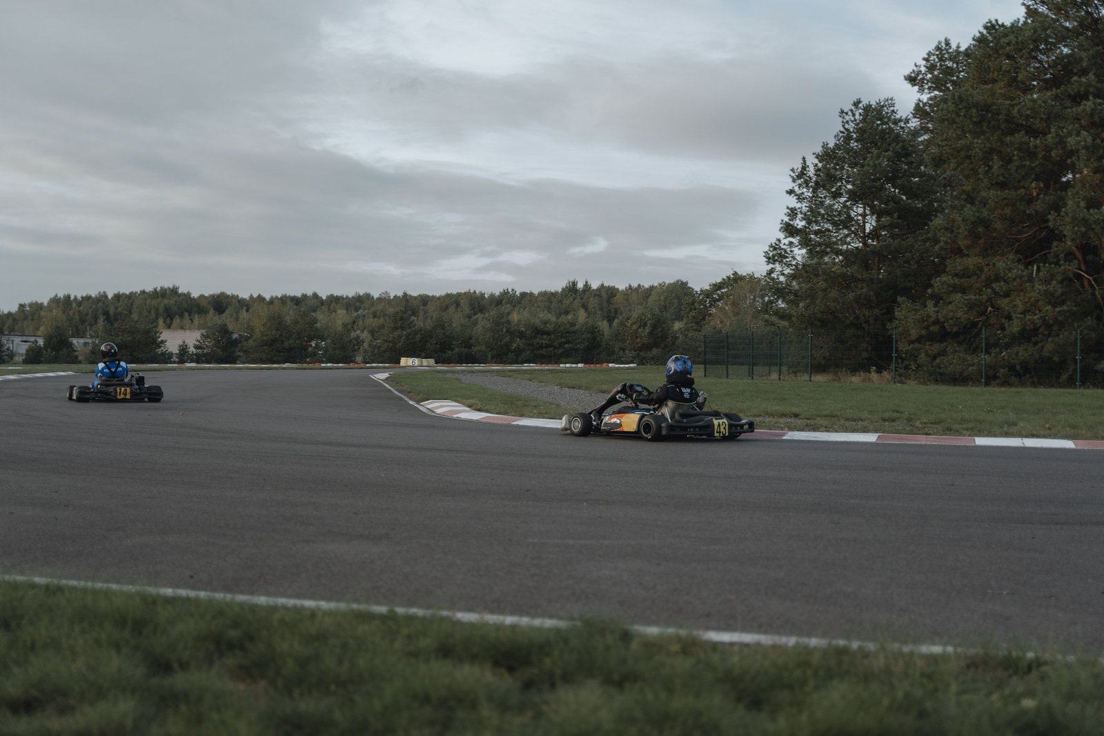 Two go-karts racing side by side on an outdoor kart circuit lined with trees